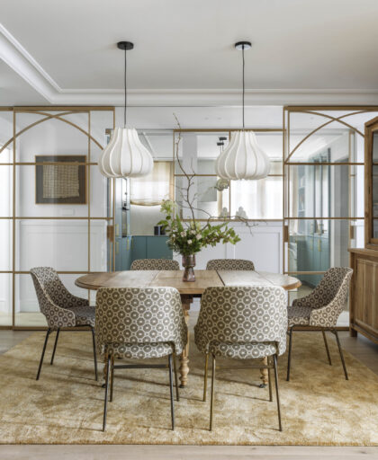Vista del comedor con mesa de madera y lámparas blancas, conectado visualmente al salón mediante cerramientos acristalados