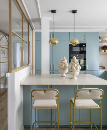 Cocina con península central, muebles verde empolvado, lámparas doradas colgantes y taburetes de estructura dorada en vivienda reformada en Getxo